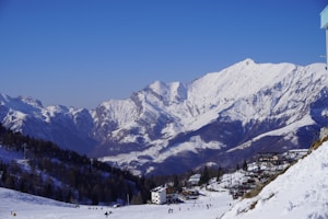 Snow-covered mountains dominate the background with a clear blue sky overhead. A snowy landscape extends into the foreground, where several small buildings and skiers can be seen on the slopes. Trees are scattered across the scene, and there is visible infrastructure for winter sports.