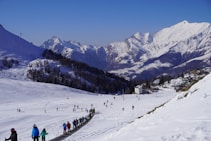 A snowy mountain landscape features a ski resort with numerous people skiing and snowboarding on the slopes. In the foreground, individuals are moving along a ski conveyor lift. The background showcases stunning mountain peaks under a clear blue sky.