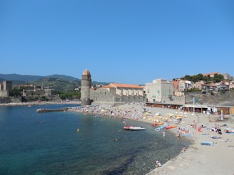 A busy beach scene with many people sunbathing and swimming in a coastal area. There is a stone tower and historic buildings with terracotta roofs nearby. The clear blue sea and sky create a vibrant backdrop, and the distant hills add depth to the landscape.