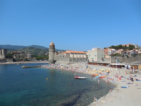 A busy beach scene with many people sunbathing and swimming in a coastal area. There is a stone tower and historic buildings with terracotta roofs nearby. The clear blue sea and sky create a vibrant backdrop, and the distant hills add depth to the landscape.