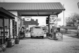 A vintage gas station scene featuring an old car parked under a canopy. Signs displaying fuel prices and brand logos from Sinclair and Valvoline are visible. The surrounding area includes wooden structures, barrels, and a landscape with bare trees in the background.