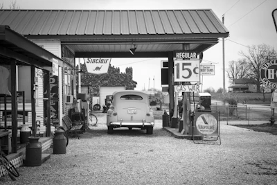 A vintage gas station scene featuring an old car parked under a canopy. Signs displaying fuel prices and brand logos from Sinclair and Valvoline are visible. The surrounding area includes wooden structures, barrels, and a landscape with bare trees in the background.