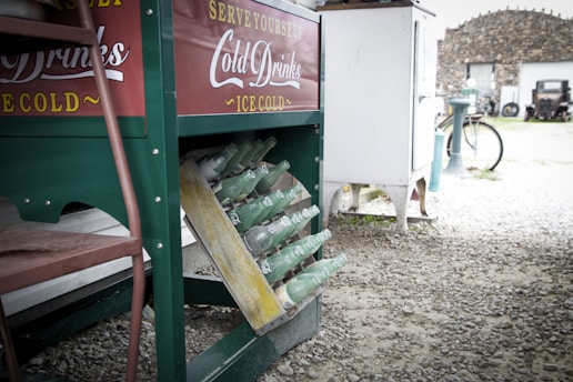 A vintage beverage cooler stands on a gravel surface, displaying rows of green glass bottles angled for easy access. The cooler has a green and red exterior with white text promoting cold drinks. A bicycle and rustic building can be seen in the background, suggesting an outdoor, antique setting.