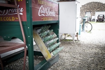 A vintage beverage cooler stands on a gravel surface, displaying rows of green glass bottles angled for easy access. The cooler has a green and red exterior with white text promoting cold drinks. A bicycle and rustic building can be seen in the background, suggesting an outdoor, antique setting.