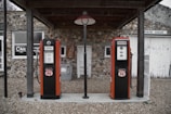 Two vintage Phillips 66 gas pumps stand under a wooden canopy in front of a rustic stone building. One pump is labeled Ethyl and the other Regular. The building features a sign for a cafe and another for Spencer Garage, with a gravel pathway leading up to it.