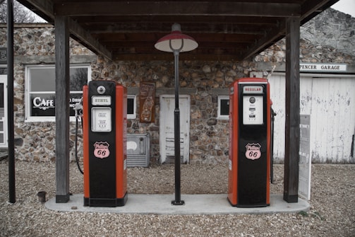 Two vintage Phillips 66 gas pumps stand under a wooden canopy in front of a rustic stone building. One pump is labeled Ethyl and the other Regular. The building features a sign for a cafe and another for Spencer Garage, with a gravel pathway leading up to it.