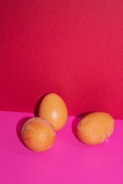 Three eggs with a light brown color are arranged on a vibrant pink surface, set against a contrasting bright red background. The eggs exhibit some uneven coloring and appear natural against the bold backdrop.