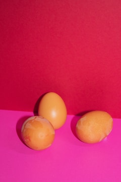 Three eggs with a light brown color are arranged on a vibrant pink surface, set against a contrasting bright red background. The eggs exhibit some uneven coloring and appear natural against the bold backdrop.