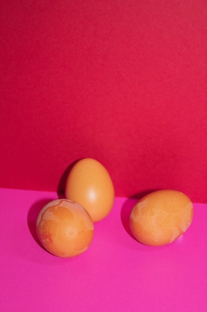 Three eggs with a light brown color are arranged on a vibrant pink surface, set against a contrasting bright red background. The eggs exhibit some uneven coloring and appear natural against the bold backdrop.