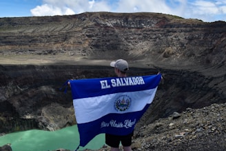 A person stands on the edge of a large volcanic crater holding a large El Salvador flag. The landscape is rugged and rocky with steep cliff walls surrounding a small, greenish lake at the bottom of the crater. The sky above is partially cloudy.