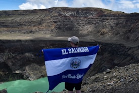 A person stands on the edge of a large volcanic crater holding a large El Salvador flag. The landscape is rugged and rocky with steep cliff walls surrounding a small, greenish lake at the bottom of the crater. The sky above is partially cloudy.
