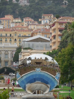 A large reflective sculpture is placed in the foreground, capturing the reflection of an ornate building, palm trees, and a busy outdoor scene. Surrounding the sculpture are lush green trees, well-maintained gardens, and people walking. In the background, an array of pastel-colored buildings with red-tiled roofs are set against a backdrop of dense greenery.