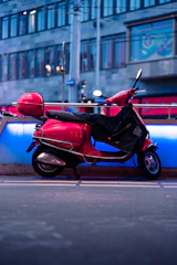 A bright red scooter is parked on a city sidewalk. It features a protective black cover partially draped over the seat. The background includes an urban street scene with blurred buildings, featuring glass windows and architectural elements. The setting appears to be in the evening, with streetlights casting a gentle glow on the scene.