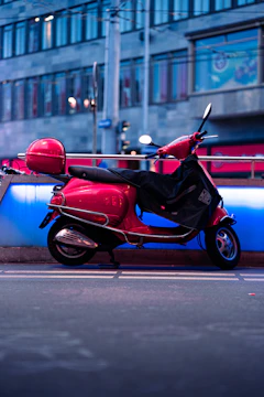 A bright red scooter is parked on a city sidewalk. It features a protective black cover partially draped over the seat. The background includes an urban street scene with blurred buildings, featuring glass windows and architectural elements. The setting appears to be in the evening, with streetlights casting a gentle glow on the scene.