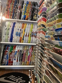 A room filled with a large collection of skateboards displayed vertically on racks, showcasing various colorful graphics and designs. The boards are organized neatly, with an assortment of vibrant colors and patterns. A Nike logo is visible on the mat on the floor, and there seems to be a warm artificial light illuminating the space.