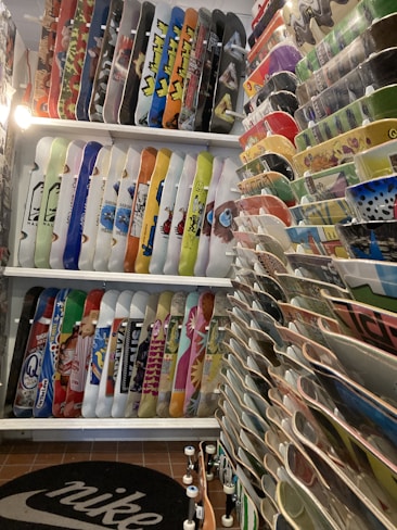 A room filled with a large collection of skateboards displayed vertically on racks, showcasing various colorful graphics and designs. The boards are organized neatly, with an assortment of vibrant colors and patterns. A Nike logo is visible on the mat on the floor, and there seems to be a warm artificial light illuminating the space.