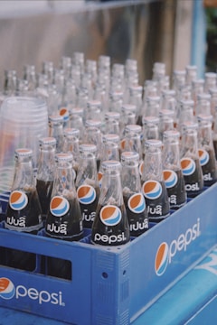 Several glass bottles of Pepsi are arranged in a blue plastic crate. The bottles have bilingual labels with the iconic Pepsi logo. Clear empty cups are stacked on the left side of the crate.
