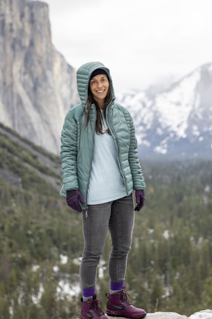 A person stands outdoors wearing a light green quilted jacket with a hood up, a light blue shirt, gray jeans, and purple gloves. They are smiling and standing against a backdrop of a forest and mountains partially covered with snow.