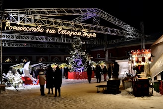 A festive outdoor night scene at a Christmas market featuring a large illuminated sign with Cyrillic text. A brightly decorated Christmas tree is central, surrounded by snow and illuminated booths. People are walking and gathering near a stall offering hot beverages. A decorative sleigh and moon seat are visible amongst snow-covered trees.
