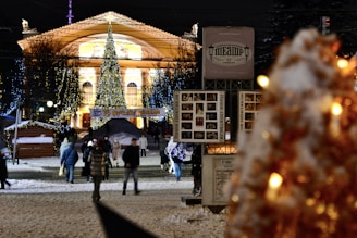 A city street at night is adorned with festive decorations. A large and brightly lit Christmas tree stands prominently in front of a grand, illuminated building. People warmly dressed in winter clothing are walking along the snow-covered street, surrounded by small wooden stalls. Colorful lights drape from the trees, adding to the festive atmosphere.