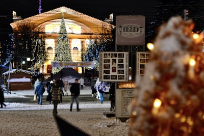 A city street at night is adorned with festive decorations. A large and brightly lit Christmas tree stands prominently in front of a grand, illuminated building. People warmly dressed in winter clothing are walking along the snow-covered street, surrounded by small wooden stalls. Colorful lights drape from the trees, adding to the festive atmosphere.