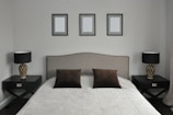 A neatly arranged bedroom features a large bed with a padded headboard. The bedding consists of a light gray quilt and two dark brown pillows. On each side of the bed are matching dark wooden nightstands, each holding a modern lamp with a patterned base and a black lampshade. Above the bed, three identical picture frames hang on the wall, each containing a blank canvas.