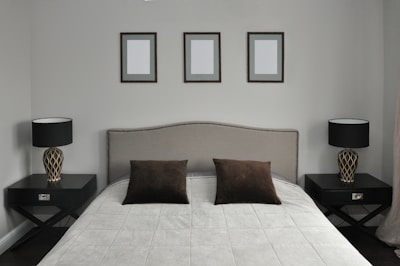 A neatly arranged bedroom features a large bed with a padded headboard. The bedding consists of a light gray quilt and two dark brown pillows. On each side of the bed are matching dark wooden nightstands, each holding a modern lamp with a patterned base and a black lampshade. Above the bed, three identical picture frames hang on the wall, each containing a blank canvas.
