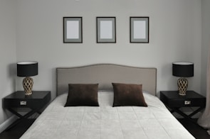 A neatly arranged bedroom features a large bed with a padded headboard. The bedding consists of a light gray quilt and two dark brown pillows. On each side of the bed are matching dark wooden nightstands, each holding a modern lamp with a patterned base and a black lampshade. Above the bed, three identical picture frames hang on the wall, each containing a blank canvas.