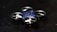 A compact, quadcopter drone with a blue body and four small propellers is positioned on a dark surface. The drone features a sleek, modern design with a central structure that is blue in color and propellers housed in circular enclosures.