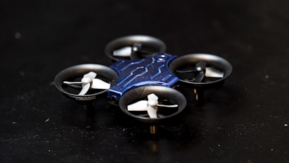 A compact, quadcopter drone with a blue body and four small propellers is positioned on a dark surface. The drone features a sleek, modern design with a central structure that is blue in color and propellers housed in circular enclosures.
