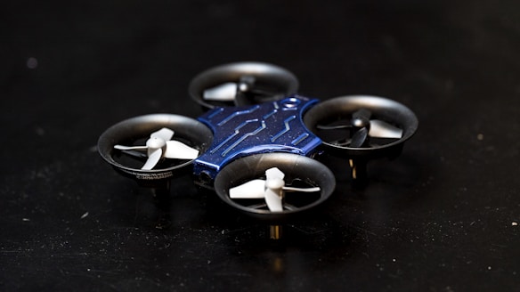 A compact, quadcopter drone with a blue body and four small propellers is positioned on a dark surface. The drone features a sleek, modern design with a central structure that is blue in color and propellers housed in circular enclosures.