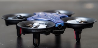 A small, blue and black drone with four propellers set against a dark background. The drone is placed on a surface, and its compact design with protective propeller guards is clearly visible.