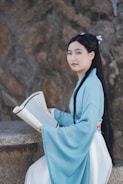 A person wearing traditional clothing is sitting at a stone table, holding an open book with printed text. The background consists of textured stone walls.