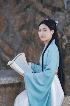 A person wearing traditional clothing is sitting at a stone table, holding an open book with printed text. The background consists of textured stone walls.