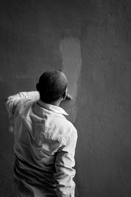 A person is seen from behind as they paint a wall. They are wearing a long-sleeved shirt and appear to be focused on applying paint evenly across the surface. The image is in black and white, emphasizing contrast and texture.