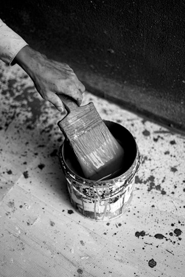 A hand holding a wide paintbrush submerged in a paint can. The surrounding area is covered in splatters of paint, suggesting an ongoing or recent painting project. The image is in black and white, highlighting the contrasts and textures.