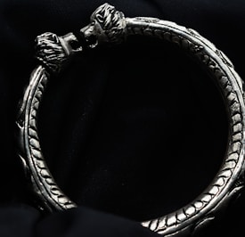 An ornate silver bracelet with intricate carvings and two lion heads facing each other at the ends. The bracelet has a textured, detailed design and sits on a dark, velvety background.
