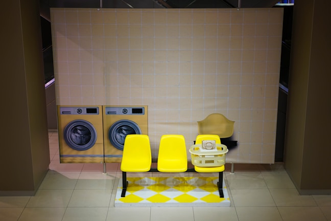 A stylized laundry setting with two front-loading washing machines depicted on a backdrop. In front of the backdrop, there is a bright yellow bench with multiple yellow chairs and a laundry basket containing a white telephone. The flooring features a yellow and white checkered pattern.