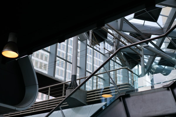 The image captures a modern architectural interior with a complex interplay of metal beams, glass elements, and geometric shapes. Natural light filters through large windows, casting reflections on the glass and metal surfaces. An industrial lamp hangs from the ceiling, adding a warm glow to the mostly cool-toned environment.