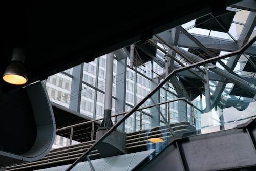 The image captures a modern architectural interior with a complex interplay of metal beams, glass elements, and geometric shapes. Natural light filters through large windows, casting reflections on the glass and metal surfaces. An industrial lamp hangs from the ceiling, adding a warm glow to the mostly cool-toned environment.
