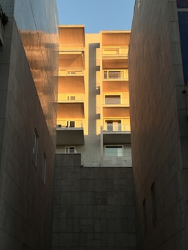 A tall apartment building with balconies is bathed in warm sunlight, creating a contrast with the shadowed walls of two adjacent buildings. The glow of the sun emphasizes the texture of the building&rsquo;s surface, and the clear blue sky provides a backdrop.