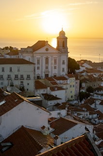 A picturesque townscape featuring a prominent white church with a bell tower, surrounded by traditional tiled-roof houses. The warm, golden glow of the sunrise or sunset casts a serene ambiance over the entire scene, with the ocean visible in the background.
