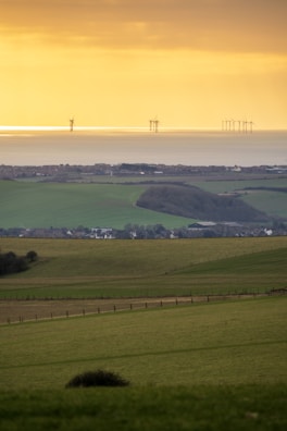 A serene landscape featuring rolling green fields descending towards a coastal area. In the background, wind turbines are visible on the horizon, standing against a golden sky at sunset or sunrise. The scene captures a mix of natural landscapes and modern renewable energy technology.