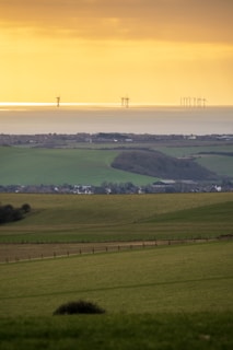 A serene landscape featuring rolling green fields descending towards a coastal area. In the background, wind turbines are visible on the horizon, standing against a golden sky at sunset or sunrise. The scene captures a mix of natural landscapes and modern renewable energy technology.