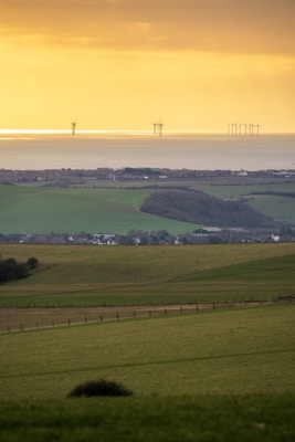 A serene landscape featuring rolling green fields descending towards a coastal area. In the background, wind turbines are visible on the horizon, standing against a golden sky at sunset or sunrise. The scene captures a mix of natural landscapes and modern renewable energy technology.