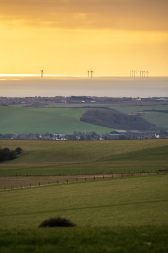 A serene landscape featuring rolling green fields descending towards a coastal area. In the background, wind turbines are visible on the horizon, standing against a golden sky at sunset or sunrise. The scene captures a mix of natural landscapes and modern renewable energy technology.