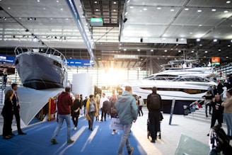 A boat exhibition featuring large, luxury yachts indoors, with several people walking around and observing. The setting includes a modern, well-lit space with high ceilings and large windows letting in natural light.