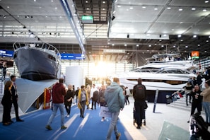 A boat exhibition featuring large, luxury yachts indoors, with several people walking around and observing. The setting includes a modern, well-lit space with high ceilings and large windows letting in natural light.