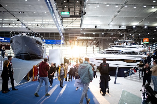 A boat exhibition featuring large, luxury yachts indoors, with several people walking around and observing. The setting includes a modern, well-lit space with high ceilings and large windows letting in natural light.