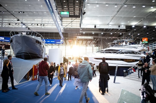 A boat exhibition featuring large, luxury yachts indoors, with several people walking around and observing. The setting includes a modern, well-lit space with high ceilings and large windows letting in natural light.